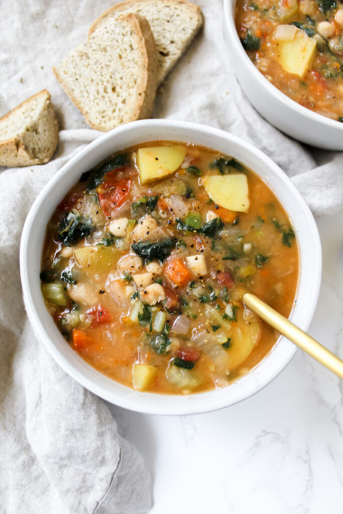 overview shot of a bowl of tuscan white bean and kale soup with a spoon in it and bread on the side