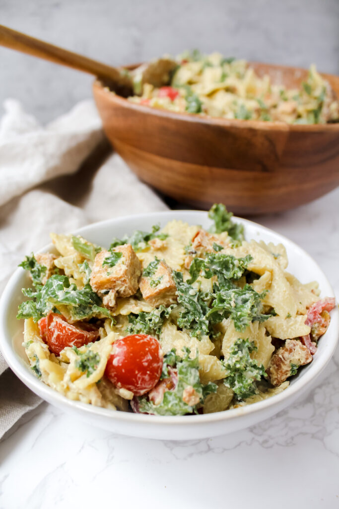 angled shot of kale caesar pasta salad with crispy tofu in white bowl and the big pasta bowl in the background 