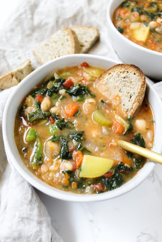 angled shot of tuscan white bean and kale soup with a slice of bread in it